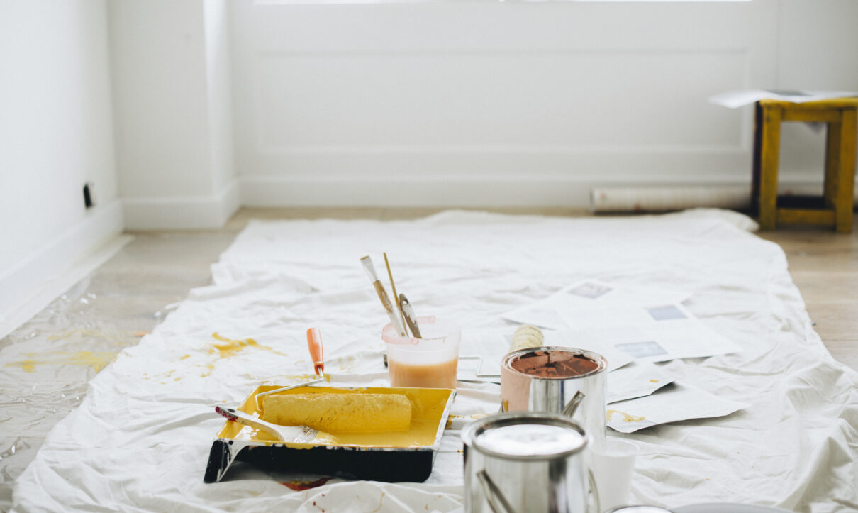 Paint cans, rollers, trays, and brushes scattered on a protective sheet in a bright room after renovation or painting work.