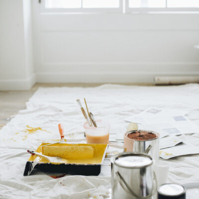 Paint cans, rollers, trays, and brushes scattered on a protective sheet in a bright room after renovation or painting work.