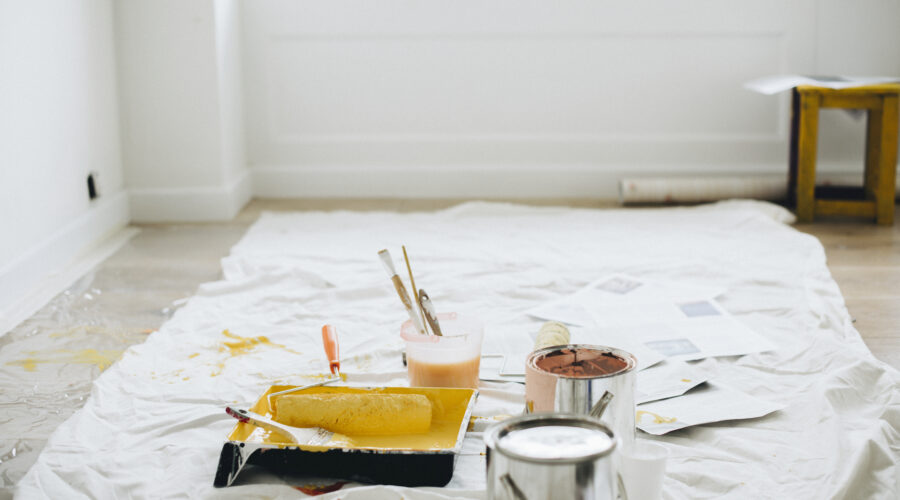 Paint cans, rollers, trays, and brushes scattered on a protective sheet in a bright room after renovation or painting work.