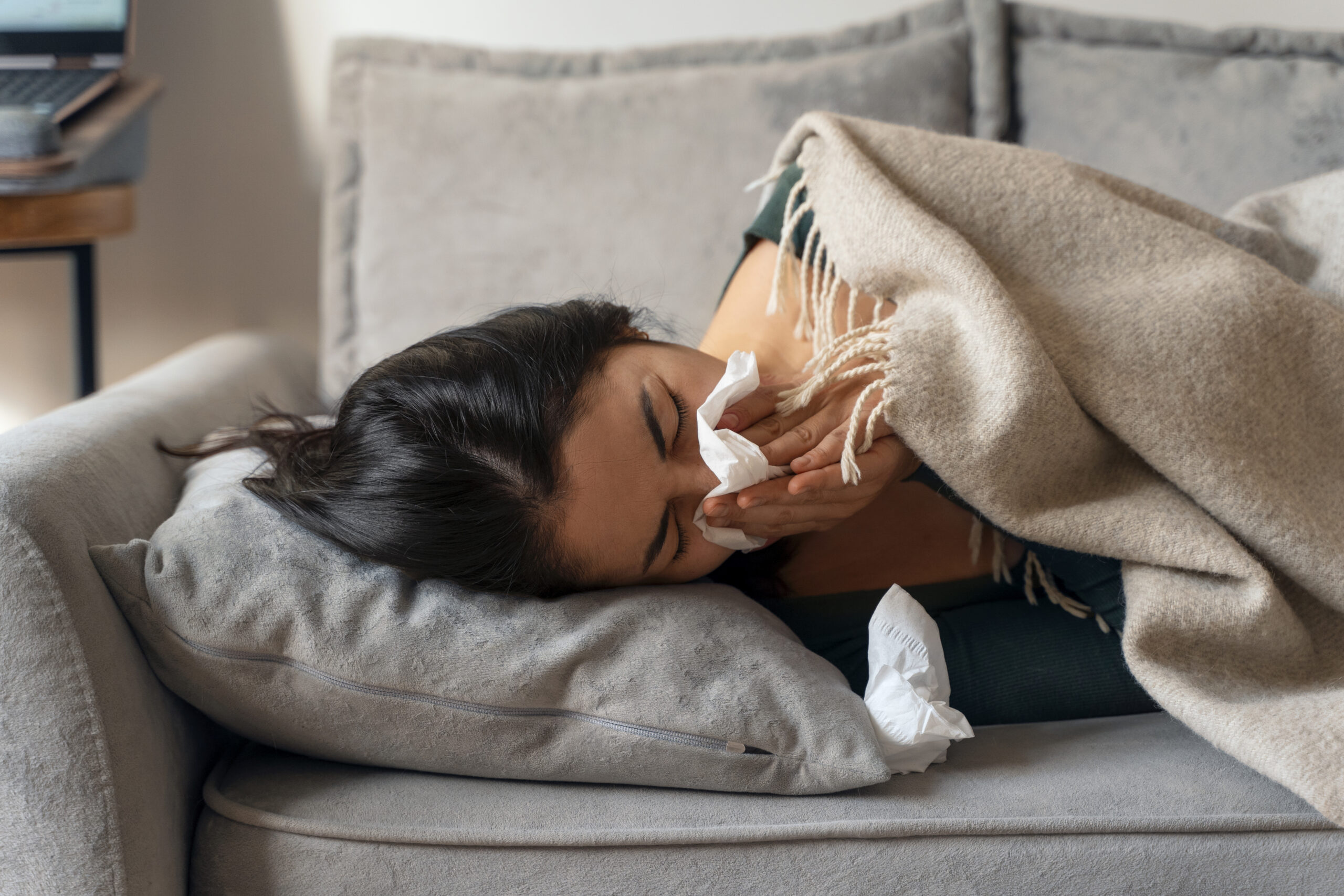 Woman lying on a couch under a blanket, blowing her nose with tissues around her, showing symptoms of illness or allergies.